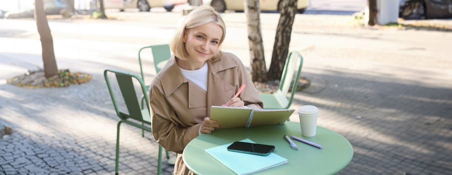 Image of young smiling woman, sitting in outdoor cafe, doing sketches, drawing art on street, spending time on coffee shop and creating art.