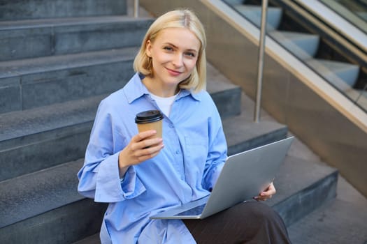 Close up portrait of stylish young female student, sitting on street stairs with laptop computer, holding coffee drink and smiling at camera.