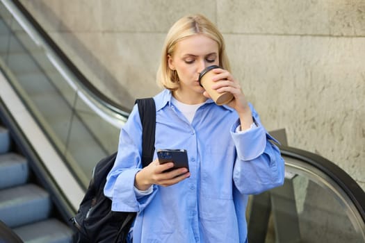 A happy, elegant businesswoman descending the escalator and holding takeaway coffee while reading important messages. A woman on the escalator with a phone.