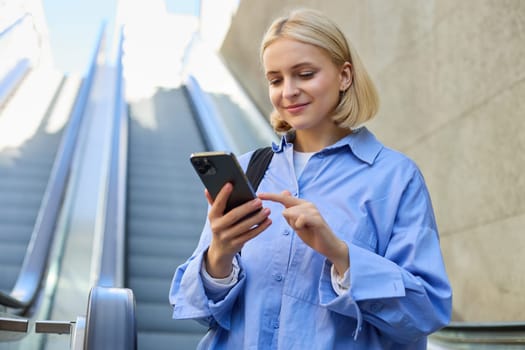 Portrait of young stylish woman, female employee, standing near escalator, using mobile phone, sending message on smartphone, tap screen, smiling and reading on telephone.