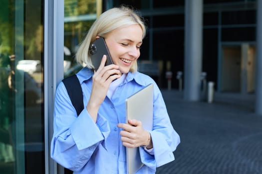 Young modern woman, female employee, standing near office building, answers phone call, talking, holding laptop and backpack, smiling while having a nice, friendly chat.
