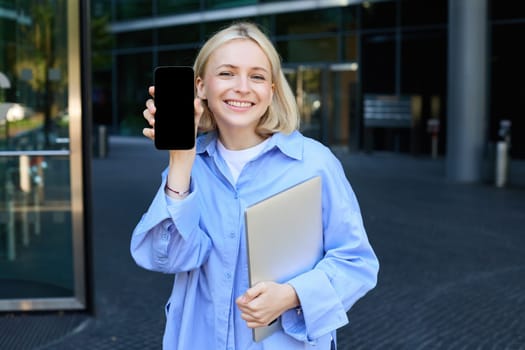 Portrait of stylish, modern young office worker, woman showing her smartphone screen, mobile phone app on display, posing near campus or office building, holding laptop.