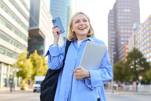 Enthusiastic blond woman, shaking smartphone in hand, holding laptop, standing on street of city centre, laughing and smiling, looking pleased, triumphing.