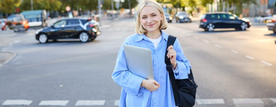 Street stylish portrait of young happy woman, blond girl with backpack and laptop, standing outdoor s near road on sunny bright day. Lifestyle concept