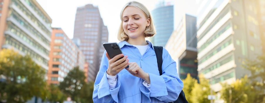 Portrait of beautiful young woman in city centre, standing in blue shirt with smartphone, checking notifications, waiting for car, order taxi on mobile phone app.