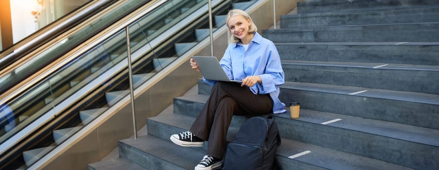 Image of young woman, student studying online, working remotely, sitting on stairs with laptop and drinking coffee in takeaway cup, elearning.