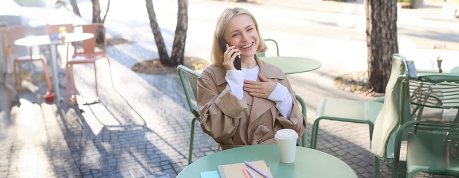 Image of stylish blond woman talking on mobile phone, drinking coffee in cafe outdoors, enjoying warm weather in city centre, answer telephone call on smartphone.