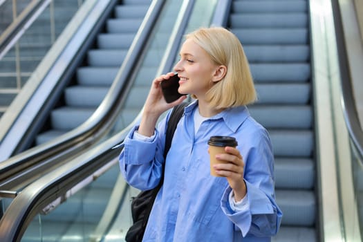 Portrait of cute young blond woman, student with cup of coffee, answers phone call, standing near escalator in city and talking, chatting with someone over the smartphone.