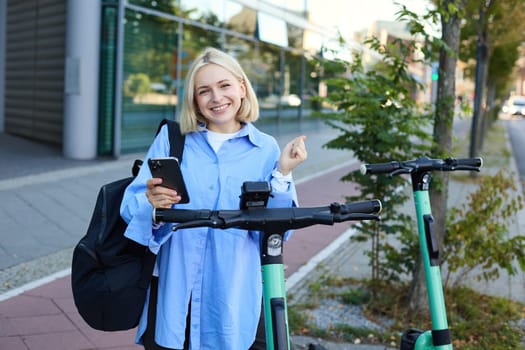 Beautiful blond girl, scans QR code on mobile phone, stands near electric street scooter to rent it, requesting a ride, smiling and looking happy.