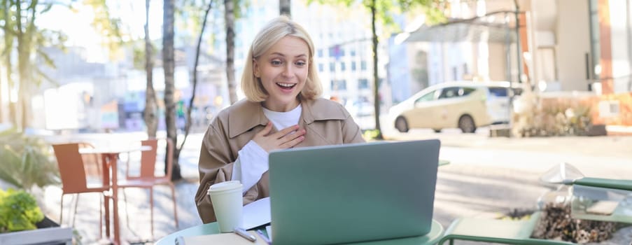 Young woman, freelancer, student doing homework in outdoor cafe, drinking her coffee on street, using laptop, connects to online lecture or doing course in internet.