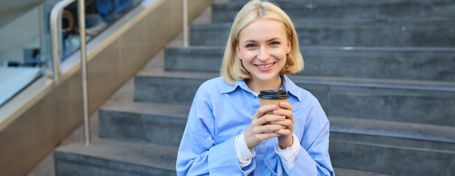 Cute young female student, woman with cup of coffee in hands, resting on stairs in city centre, sitting and drinking cappuccino, smiling happily.