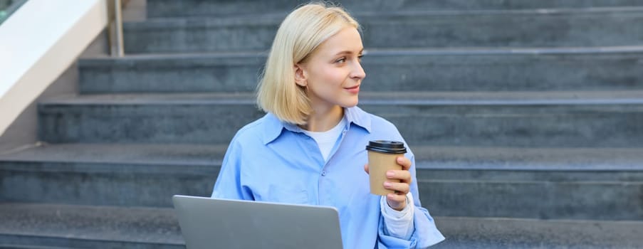 Young woman sitting with cup of coffee and laptop on stairs outdoors, working remotely, freelancing, connecting to public wifi in city to study online.