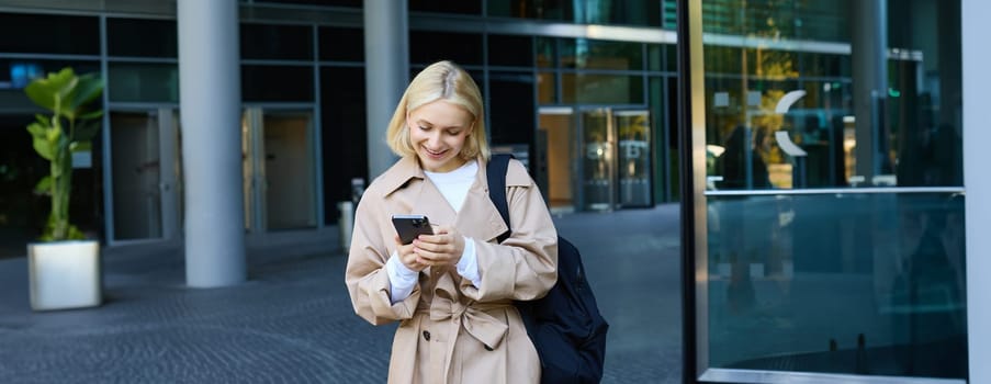 Street photo of young blond woman with smartphone, walking in city, waiting for someone, order taxi on mobile application.