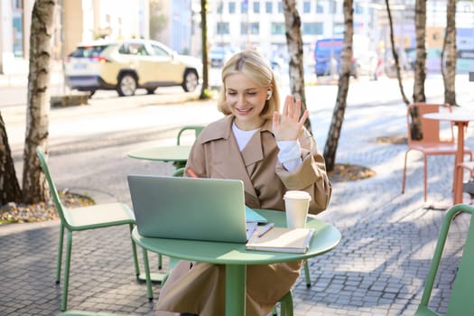 Cheerful girl with laptop, saying hello, connects to online meeting, waving hand at camera, chatting with someone via internet, sitting in outdoor coffee shop.