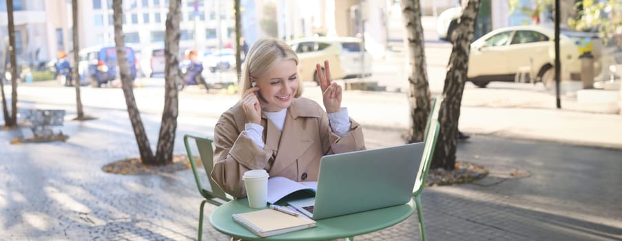Image of smiling, beautiful young woman in wireless headphones, connects to online meeting, video chatting in outdoor coffee shop, showing peace sign, using laptop, studying remotely.