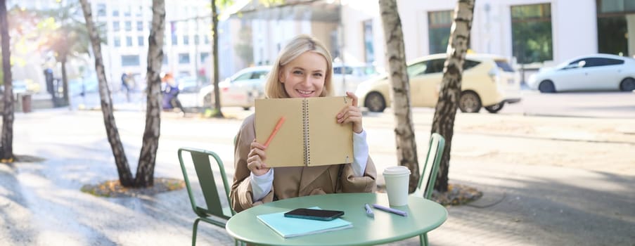 Portrait of young woman sitting in outdoor cafe, hiding behind journal, holding notebook in hands and smiling.