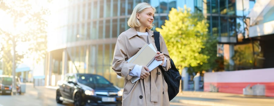 College lifestyle and student concept. Smiling blonde woman on street, sunny day, skyscraper behind her, girl holding documents, notebooks and backpack.