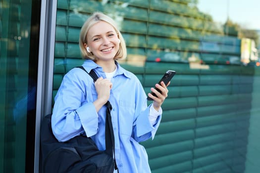 Lifestyle portrait of smiling young female model, student with backpack, waiting for someone on street, standing outdoors with smartphone.