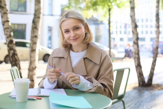 Portrait of carefree, blond cute woman working with documents, drinking takeaway coffee and sitting in cafe outdoors with documents, writing essay, student doing homework.