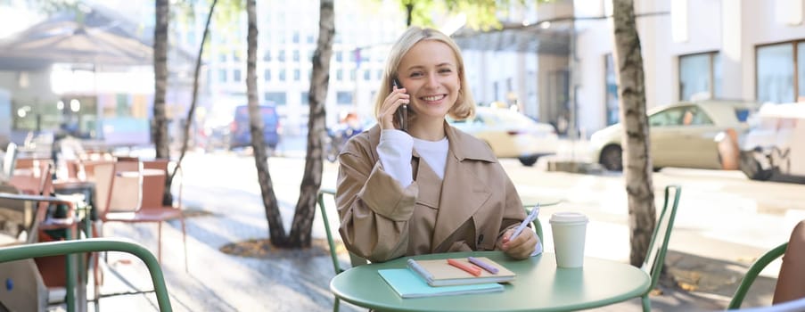 Friendly young woman in trench, sitting outdoors on sunny day, spending time in cafe, answer phone call, talking on mobile.