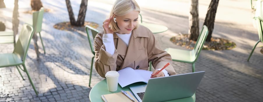 Portrait of young stylish woman, student in outdoor coffee shop, using laptop and making notes, wearing wireless headphones, listening to online course.