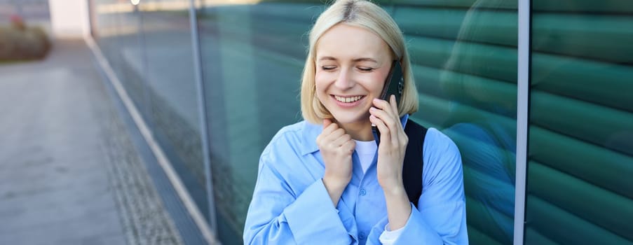 Lifestyle portrait of happy blond woman, chatting over the phone, answers a call, laughing and smiling, standing on street and looking cheerful.