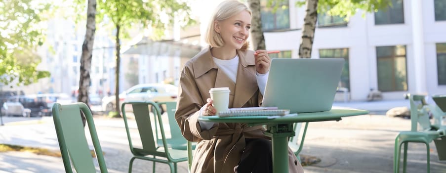 Image of young blond modern woman, sitting with laptop outside in cafe, drinking coffee drink and working on project, using computer.