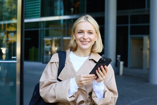 Lifestyle portrait of young beautiful woman, waiting for someone on street, holding smartphone, using mobile phone, ordering taxi in application.