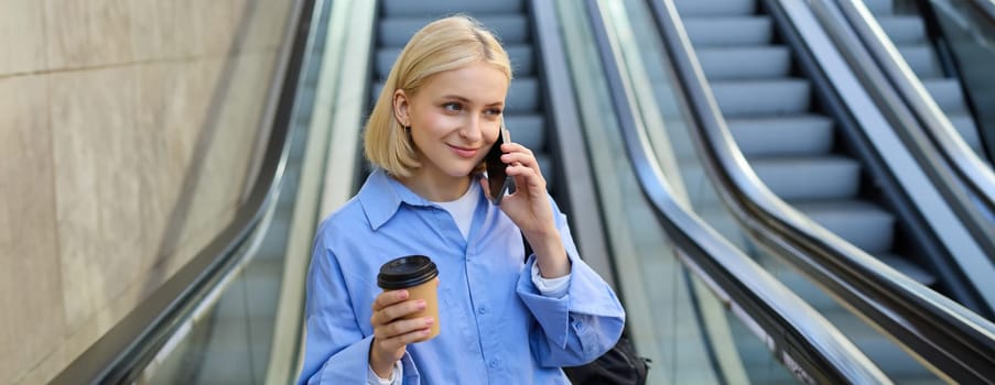 Portrait of cute young blond woman, student with cup of coffee, answers phone call, standing near escalator in city and talking, chatting with someone over the smartphone.