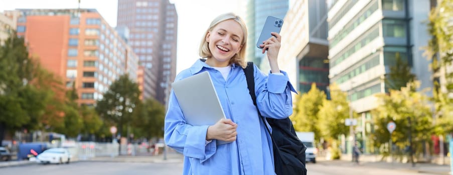 Enthusiastic blond woman, shaking smartphone in hand, holding laptop, standing on street of city centre, laughing and smiling, looking pleased, triumphing.