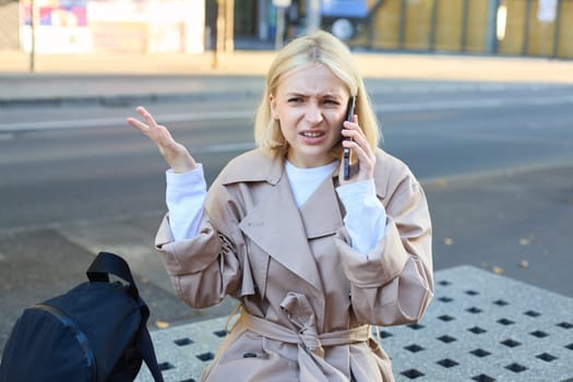 Frustrated young woman sitting on bench with smartphone, answer phone call, talking on mobile and shrugging, looking disappointed, spending time outside.