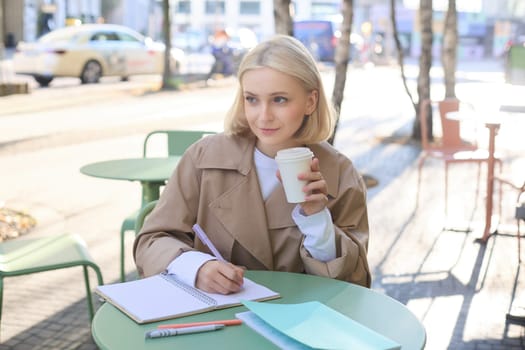 Portrait of beautiful blond girl, college student writing in notebook, doing homework in outdoor cafe, drinking coffee, smiling and looking at the street.
