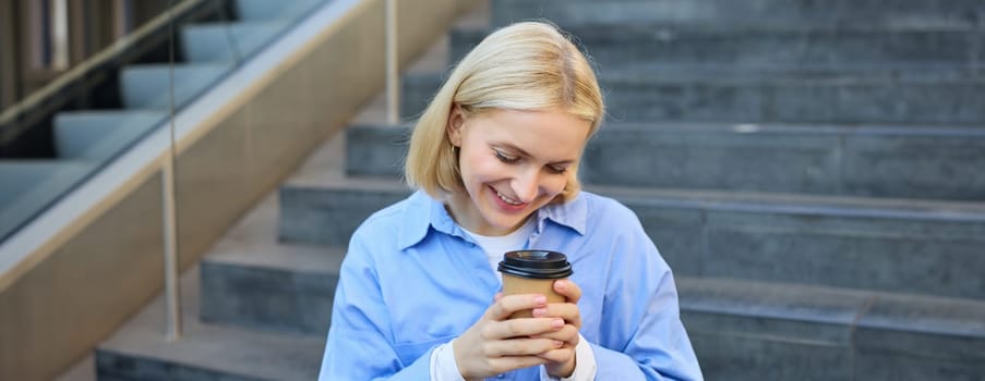 Cute young female student, woman with cup of coffee in hands, resting on stairs in city centre, sitting and drinking cappuccino, smiling happily.