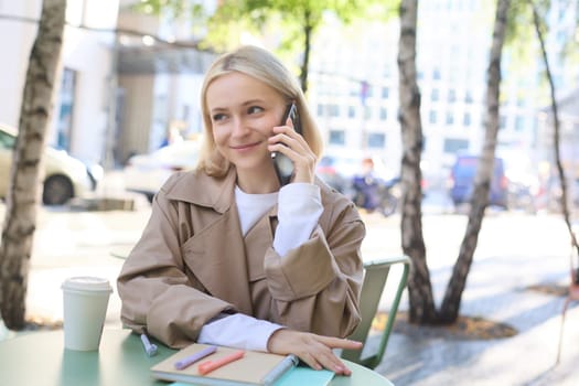 Close up portrait of smiling blond woman, sitting at table outside cafe, talking on mobile phone, having happy, lively chat.