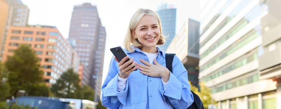 Close up portrait of stylish young blonde woman, standing on street, checking her mobile phone, using smartphone app to get around town, looking at online map for directions.