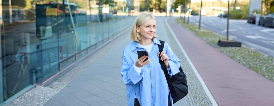 Portrait of blond young woman, using navigation map app on smartphone, walking along street, using directions on phone, holding backpack.