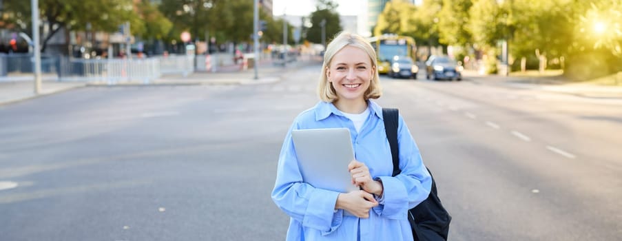 Street stylish portrait of young happy woman, blond girl with backpack and laptop, standing outdoor s near road on sunny bright day. Lifestyle concept