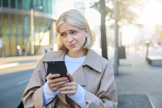 Portrait of young blond woman standing on street, has unsure, doubtful face expression, using mobile phone, showing reluctance.