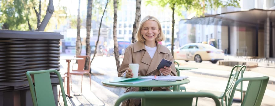 Happy beautiful young woman enjoying the sun outdoors, sitting in city centre cafe, drinking coffee, holding smartphone and laughing, smiling with eyes closed.