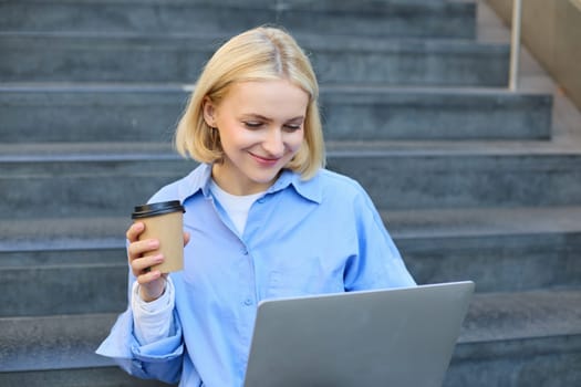 Smiling young urban woman, student working on laptop, sitting outdoors on stairs, drinking coffee, studying, e-learning on fresh air.