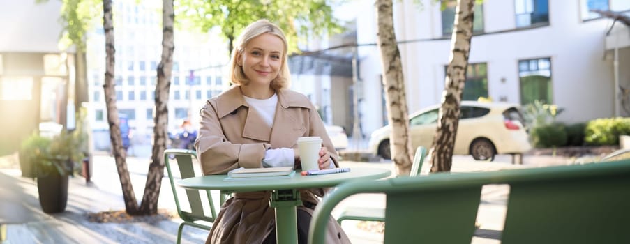 Portrait of stylish modern woman, sitting in an outdoor cafe, smiling and drinking coffee from takeaway cup, wearing trench coat.