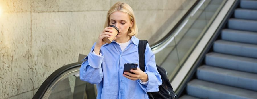 A happy, elegant businesswoman descending the escalator and holding takeaway coffee while reading important messages. A woman on the escalator with a phone.