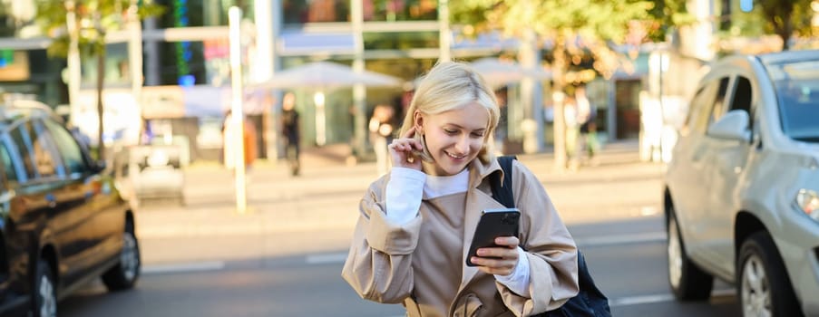 Modern young woman on street, reading message on mobile phone, using smartphone, standing near busy road and smiling.