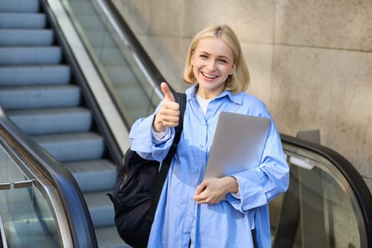 Friendly, smiling young woman, college student with laptop and backpack, using escalator, showing thumbs up in approval, recommending product or company.