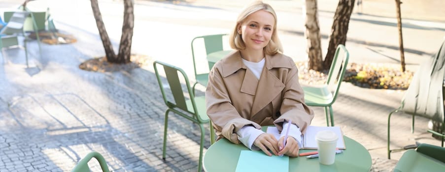 Image of young female entrepreneur, businesswoman drinking coffee in outdoor cafe and writing in notebook, making notes, working on documents.