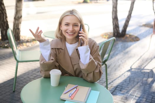 Image of stylish blond woman talking on mobile phone, drinking coffee in cafe outdoors, enjoying warm weather in city centre, answer telephone call on smartphone.