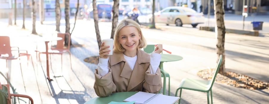 Portrait of beautiful blond woman, smiling as she is drinking coffee in outdoor cafe, writing notes in notebook, doing homework, preparing material for project.