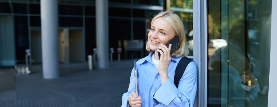 Close up portrait of modern young woman, student standing near building in city centre, talking on mobile phone, having chat on smartphone and smiling.