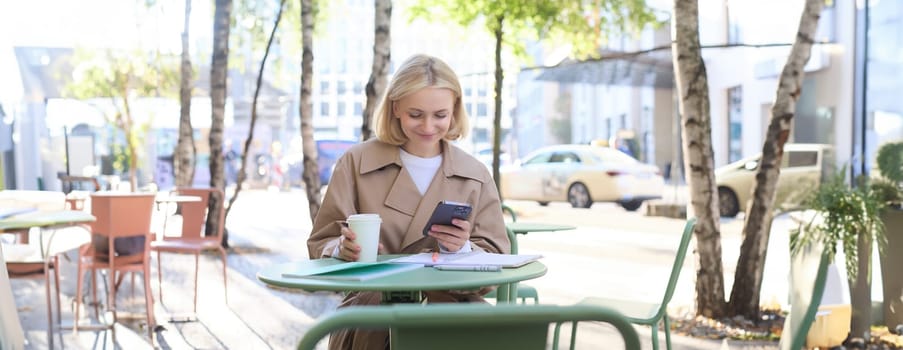 Image of young woman drinking coffee, sitting in outdoor cafe with smartphone, sending message, reading notification on mobile phone and smiling.