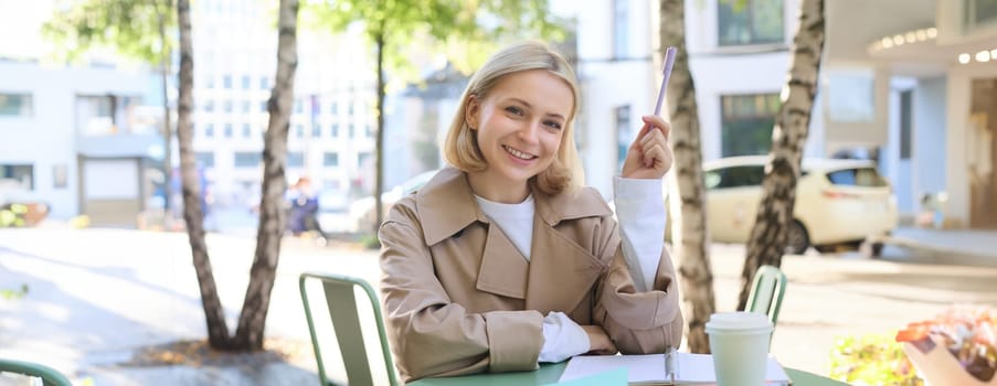 Portrait of young female student, working on her documents, doing homework outdoors in cafe, drinking coffee and writing essay, holding marker, smiling at camera.